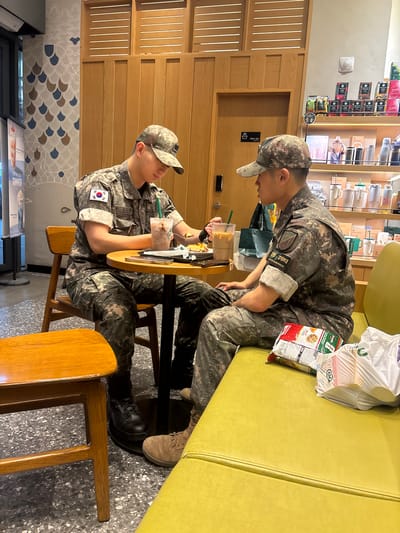 Two individuals in South Korean military camouflage uniforms are seated at a table in what appears to be a cafe. One person is leaning over the table, holding a drink with a straw, and looking at something on the table. The other person is seated opposite, looking towards the first individual. They are both wearing camouflage baseball caps. On the table, there are drinks and what looks like snacks. Behind them, shelves are stocked with various items, possibly merchandise or food products. A yellow couch is visible in the background. Visible text includes "최전방 경계부대" (Frontline Guard Force), "PARTNER SPACE", "Dontas", and "CU". The setting seems to be indoors, with wood paneling on the walls and a patterned wallpaper. The lighting suggests it is daytime.
