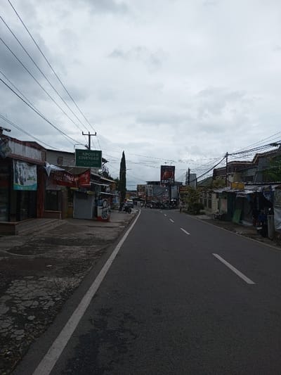 The image displays a street view identified as being in the city of Melakudikadu, India. The sky is overcast with a uniform grey cloud cover. A two-lane asphalt road, marked with a continuous white line along its left edge and intermittent dashed white lines in its center, stretches away from the viewer.
On the left side of the road, a cracked concrete sidewalk fronts a row of one to two-story commercial buildings. A prominent green sign, mounted on a pole, reads "APOTEK CINENGAN." Below it, a red and white banner states "MAU KEB PAKAI SUTRA." Two manual fuel dispensers, bearing labels such as Coca-Cola and Kopi ABC, are visible near the sidewalk. Overhead, multiple black electrical and communication wires extend across the street, supported by a wooden utility pole. A solitary individual is partially visible near the fuel pumps on the left.
The right side of the street also features a series of one to two-story buildings, some appearing as shops and others residential. Several motorcycles are parked along this side. Further down the street, another utility pole is heavily laden with numerous entangled wires. A large billboard displaying an advertisement is present in the mid-distance on the right.
The overall scene depicts an active urban environment with a mix of commercial establishments. Human and vehicular activity appears minimal, with no significant movement observed. Signs visible, specifically "APOTEK CINENGAN" and "MAU KEB PAKAI SUTRA," are in Indonesian.