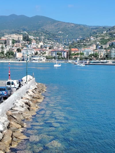 A bright, sunny day unfolds over a coastal city in Sanremo, Italy, where the azure Mediterranean Sea meets a rocky shoreline. In the foreground, a sturdy concrete pier juts out into the water, lined with large, uneven rocks that form a natural barrier against the waves. Parked along the pier are a white van, a motorcycle, and at least two cars. A red beacon with a solar panel sits atop a pole near the edge of the pier, marking its presence. The clear water reveals the rocky seabed beneath, with sunlight creating dappled patterns on the submerged stones.

Further out in the bay, two sailboats glide across the calm water, their white sails stark against the deep blue. The city of Sanremo sprawls across the hills in the background, a tapestry of buildings with terracotta roofs and white facades, interspersed with lush greenery. Tall palm trees dot the landscape, hinting at a Mediterranean climate. In the distance, a modern bridge spans a valley, and the silhouettes of mountains rise majestically against a clear blue sky. The scene evokes a sense of tranquil coastal life, a vibrant city basking in the Mediterranean sun.