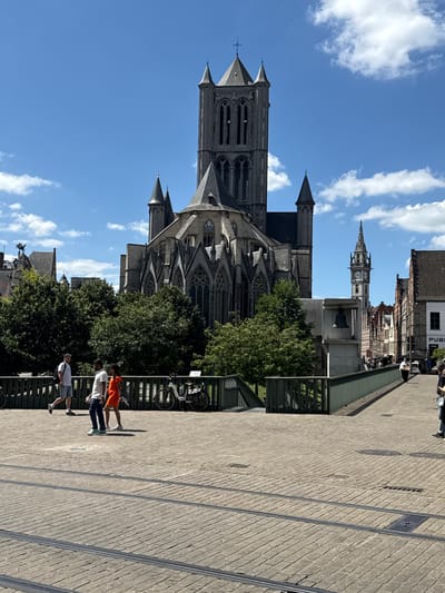 A vertical medium shot captures a scene in Ghent, Belgium, on a sunny day with scattered clouds. The focal point is the imposing Gothic architecture of a large church, likely St. Bavo's Cathedral, with its prominent towers and intricate stone carvings. Adjacent to the church, a tall, slender clock tower with visible clock faces stands out against the blue sky.
In the foreground, a paved walkway made of grey bricks runs diagonally across the frame. A group of four people are walking on this path. The first person from the right wears a white t-shirt and dark shorts. Next to them, a person in an orange top and dark pants walks ahead. Following them are two more individuals, one in a white t-shirt and jeans, and another in a grey t-shirt and shorts. They appear to be casually strolling.
To the left of the walkway, lush green trees partially obscure the view of the church. A green metal fence or railing separates the walkway from the area with the trees and church. A bicycle is visible leaning against the railing.
The lighting suggests it is daytime, likely mid-morning or early afternoon, given the clear blue sky and the shadows cast by the people. The overall atmosphere seems peaceful and pleasant, characteristic of a tourist-friendly location. No discernible text is visible in the image.