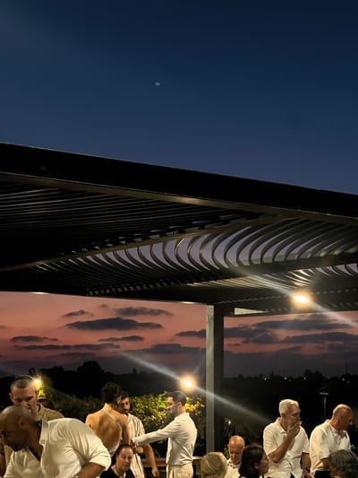 At dusk, a gathering of people is taking place outdoors in Beit Hanan, Israel, under a pergola structure. The sky displays a gradient of dark blue at the top transitioning to hues of pink and orange, with scattered clouds. Several individuals, primarily men dressed in white shirts and some in kippahs, are visible. Some are standing and conversing, while others are seated at tables. One man is seen smoking. The setting appears to be a social event, possibly a celebration or a meal, with ambient lighting from the fixtures on the pergola. The background shows a dark silhouette of trees and vegetation against the twilight sky.