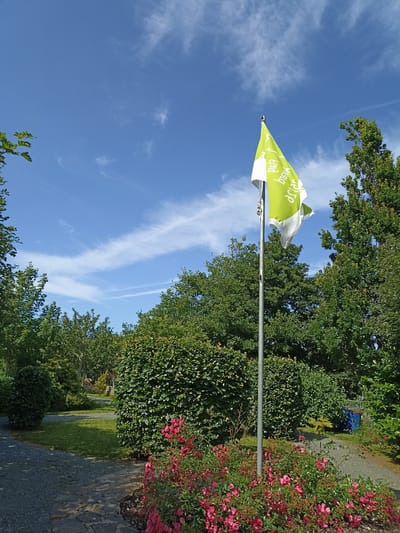A green and white flag with text is planted on a metal pole in a garden. The flag is illuminated by the sun. Behind the flag, there are lush green trees and bushes. In the foreground, there is a bed of pink roses. The ground is covered with gravel and stone paths, and there is a patch of green grass. The sky above is a clear blue with scattered white clouds. The image appears to have been taken in Dungloe, Ireland, based on the information provided.