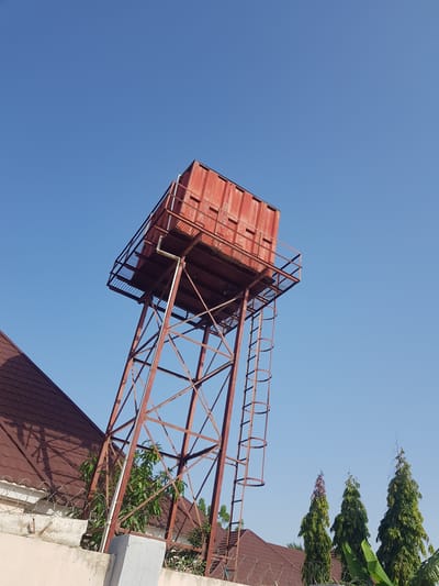 This media file captures a low-angle view of a red, rusty water tower against a clear blue sky. The water tower is supported by a metal lattice structure. A ladder with a protective railing leads up to the platform of the tank. The tank itself appears to be a large, rectangular container. The scene is set outdoors, with glimpses of a tiled roof and lush green trees in the background. Barbed wire is visible along the edge of what appears to be a wall or fence in the foreground. The clear sky suggests it is daytime, likely a sunny day. The setting appears to be in Palade, Nigeria. No people are visible in the image. There is no visible text.