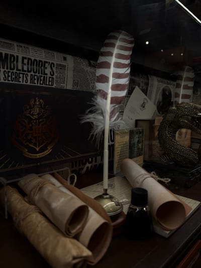 A collector's display features a quill pen with red and white stripes nestled in a quill stand near rolled parchment, an inkwell, and a golden dragon claw. Scattered newspapers with articles titled "Dumbledore's Secrets Revealed" and Hogwarts crests are present. A card labeled "E4999" rests among the items, showcasing a vignette of the Harry Potter universe.