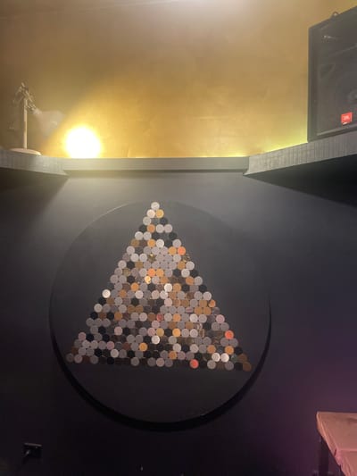 The image depicts a wall art installation resembling a triangle made of numerous circular metallic objects. This artwork is mounted on a dark grey circular background against a dark grey wall. Above the wall art, a shelf juts out, supporting a white desk lamp with a flexible arm on the left and a black JBL speaker on the right. The wall above the shelf is painted in a textured golden hue. The scene appears to be indoors, possibly in a bar or entertainment venue, with dim lighting and a hint of purple light in the lower right corner. The overall impression is one of artistic display within a dimly lit interior. The image does not contain any people.