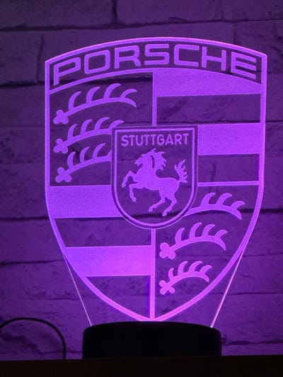The image shows a purple, illuminated Porsche logo. The logo includes the word "PORSCHE" along the left side and the Stuttgart coat of arms in the center, depicting a horse. The background appears to be a brick wall. There is no indication of Marche-en-Famenne, Belgium in the provided image.