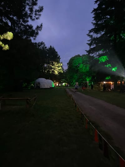 The image captures an outdoor nighttime scene within the Sremski Karlovci Municipality, Serbia. A wide dirt path extends from the foreground into the background, curving slightly to the right. The sky above is a dark blue-purple, indicative of late twilight or early night.
Dense, dark trees border the scene on both sides, with some branches silhouetted against the sky and others vividly illuminated by artificial lights. On the left side of the frame, a large, white geodesic dome or spherical tent structure is visible, with its base bathed in green light. Further into the background, a series of smaller structures, possibly stalls or temporary buildings, are illuminated by warm, yellow string lights. The foliage of several large trees in the midground and right side of the image is intensely illuminated by bright green spotlights, creating distinct glowing effects and visible light beams.
Along the right edge of the dirt path in the foreground, a low demarcation line is marked by a white rope strung with small, alternating red and dark-colored triangular flags. Multiple individuals are present throughout the scene. Two figures are visible on the dirt path in the midground, one appearing to wear a light-colored top. Additional people are discernible near the illuminated structures in the background. The ground consists of dark grass flanking the central dirt path. The scene is dominated by artificial lighting, which casts vibrant green hues on the vegetation and structures, contrasting with the overall darkness and the warm tones of scattered string lights.