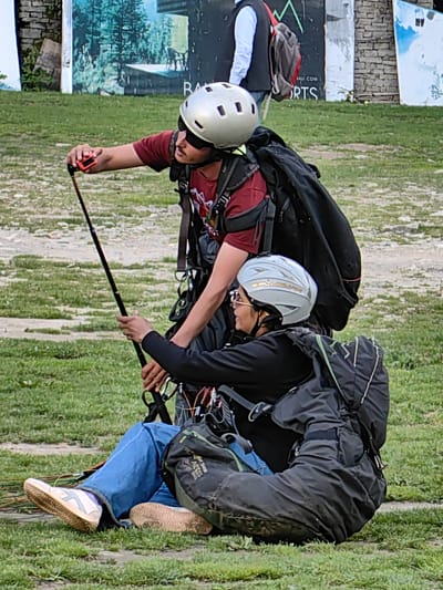 This image captures two individuals in Solang, India, likely preparing for or having just completed paragliding. A man, wearing a maroon t-shirt, sunglasses, and a silver helmet, is holding a selfie stick with a small orange device attached to its end. He is looking towards the right side of the frame, and his left arm is extended, holding the stick. To his right and slightly in front, a woman is seated on the grass, also wearing a silver helmet and sunglasses. She is wearing blue jeans and a black jacket and appears to be interacting with the man or focusing on the selfie stick. Both individuals are equipped with large backpacks, typical of paragliding gear, and are surrounded by a grassy field. In the background, a large advertisement or mural depicting a forest scene and buildings is visible. The ground is a mix of grass and dirt.