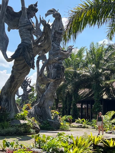 The image depicts a sculpture in a garden setting in Kedewatan, Indonesia. The sculpture appears to be made of gray stone and depicts figures with intricate details. Lush greenery surrounds the sculpture, including palm trees and other tropical plants. A woman stands on a paved path that runs through the garden. The sky is blue with scattered clouds. A building with a dark roof is visible in the background.