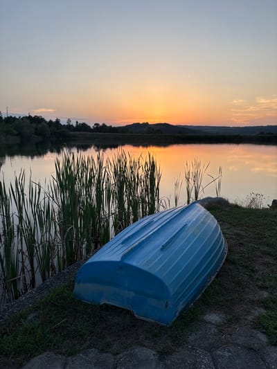 A blue boat is overturned on the grassy bank of a calm lake during sunset. The sky is painted with hues of orange and yellow, reflecting on the water's surface. Tall reeds grow along the water's edge. The scene is peaceful and serene, set in Skalsko, Bulgaria.