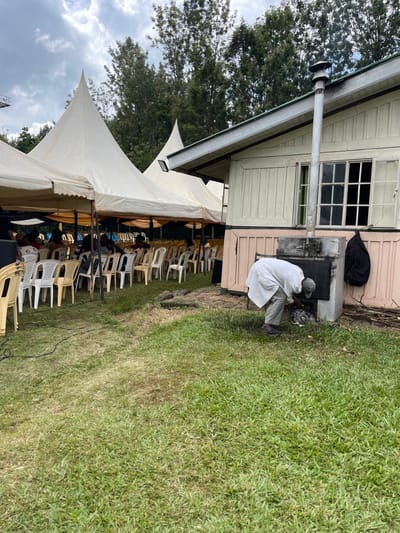 This image depicts an outdoor scene in Makuyu, Kenya, likely during a daytime event under an overcast sky.
In the midground, a light-colored building, possibly a house or a shed, features multi-paned windows and a metal chimney pipe extending from its roof. Adjacent to this building, a person wearing a long white robe or coat and a light-colored hat is crouched down, tending to a dark-colored, stove-like structure built into the ground next to the building. This structure appears to be an outdoor oven or cooktop, with visible embers or fire being managed by the individual. A black bag rests against the building near this outdoor cooking setup.
To the left of the building and extending into the foreground, a large white tent with pointed peaks is erected on a grassy area. Numerous empty plastic chairs, predominantly white and beige, are arranged in neat rows both under and outside the tent, suggesting a gathering or event is either about to begin or has recently concluded.
The ground is covered in vibrant green grass, with a dark cable or wire running across it in the lower right. In the background on the left, tall trees with dense green foliage are visible against the backdrop of a cloudy, overcast sky. The scene conveys an atmosphere of preparation for an outdoor communal activity. No discernible text is visible within the image.