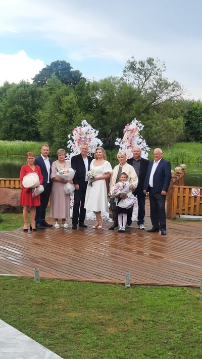 A group of nine people, including the bride and groom, are posing for a photograph on a wooden deck near a body of water. The bride is in a white dress, holding a bouquet of white flowers. The groom is in a black suit. The group includes adults and one young child. Behind them is a floral wedding arch and lush greenery. The scene appears to be outdoors, possibly at a wedding venue in the Жодзішкаўскі сельскі Савет, Belarus region, given the natural landscape. The wooden deck is wet, suggesting recent rain.