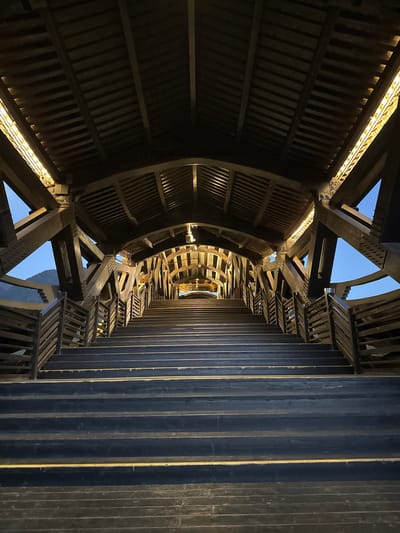 An upward-angled shot captures the interior of a wooden structure, possibly a tower or observation point, located in Zhuoshui, China. The structure features a circular, slatted wall on the left, contrasting with a series of vertical wooden planks to the right. Interconnecting beams and supports are visible throughout the frame, creating a complex network of geometric shapes. Gaps in the structure reveal glimpses of a pale blue sky. Embedded lighting enhances the architectural detail and depth of the image.