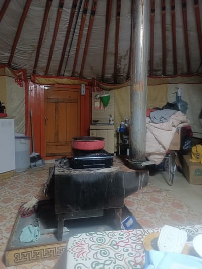 The image depicts the interior of a ger, likely in Ulaanbaatar, Mongolia, given the context. A central wood-burning stove, made of dark metal, dominates the foreground. On top of the stove sits a red pot. A metal flue pipe rises vertically from the stove, extending towards the ger's ceiling.
To the left of the stove, a white appliance, possibly a refrigerator, is partially visible. Next to it is a large, translucent plastic container filled with water, topped with a blue lid. A broom leans against the wall near a closed wooden door painted orange.
The walls of the ger are covered with fabric, adorned with decorative patterns in red and gold. The ceiling is supported by wooden poles.
To the right of the stove, a shelf unit holds various items, including cleaning supplies and a large blue water dispenser. A pile of folded blankets and clothing is stacked on a wheeled cart behind the stove and water dispenser. In the far right background, cardboard boxes are visible.
The floor is covered with a patterned rug. A light blue cloth is placed on a metal tray in front of the stove. A pair of work gloves is also visible on the tray. A portion of a table with a patterned tablecloth is in the lower right corner of the image. The overall scene suggests a lived-in space with domestic activities.