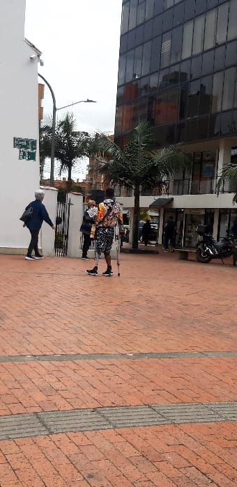 The image displays an urban pedestrian area at the intersection of Diagonal 79B and Carrera 10 in Bogota, Capital District, Colombia, under an overcast sky. The ground is covered with reddish-brown brick pavers, intersected by two horizontal grey concrete strips, one of which exhibits a grooved texture consistent with tactile paving.

A male individual, positioned in the mid-ground, is ambulating rightward, supported by two underarm crutches. He possesses a black, blade-style prosthetic on his left leg, and wears black athletic shoes on both feet. His attire consists of a brightly colored, multi-patterned long-sleeved top and black and white patterned shorts.

To the left of this central figure, a person with grey hair, dressed in dark clothing and carrying a black shoulder bag, walks towards the right. Further back and slightly to the left, another person in a dark jacket stands near a black metal gate, which provides access to an area between buildings.

The background on the right is dominated by a tall, modern building featuring a dark glass facade that reflects orange-toned elements. Its ground floor hosts multiple commercial storefronts. On the left, a white two-story building with a brown roof section displays a green street sign identifying "Dg. 79 B" and "Cra. 10". Several palm trees are scattered throughout the scene, interspersed between structures. A black motorcycle is parked on the far right, adjacent to a stone bench. Other indistinct human figures are visible further in the background near the glass building. No direct interactions between individuals are discernible.