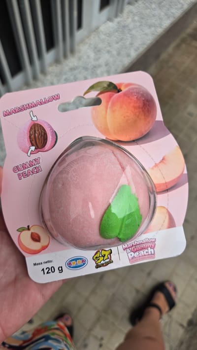The image shows a close-up of a peach-shaped gummy candy packaged in plastic. The candy is pink with a green leaf detail and is presented in pink packaging with illustrations of peaches. Text on the packaging indicates "MARSHMALLOW & GUMMY PEACH" and "WHITE PEACH FLAVOUR". Below the candy, the weight "120 g" is displayed along with brand logos. A person's hand is holding the packaging, and a portion of their legs and feet wearing sandals are visible at the bottom of the frame. The background suggests an outdoor setting with a textured floor and vertical structures. This scene appears to be in Guía de Isora, Spain, possibly in a commercial area or street.