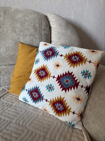 A medium shot captures two decorative pillows resting on a beige couch. The pillow in the foreground is white with a repeating Southwestern-inspired pattern featuring diamond shapes in red, blue, and yellow. Behind it, to the left, is a plain mustard-yellow pillow. The couch appears to be made of a textured fabric, and a textured beige blanket is draped over the seat. The background shows the textured wall of a room. The scene is static and peaceful.