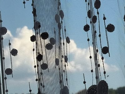 The image shows a close-up view of a sheer curtain with a decorative pattern of dark circles and vertical lines. The curtain is hanging in front of a window, and through it, a bright sky with fluffy white clouds can be seen. The light suggests it is daytime. The pattern on the curtain creates a visual layer between the viewer and the sky. The overall impression is one of a slightly obscured, serene outdoor view, possibly from an indoor setting. There is no visible text in the image.
