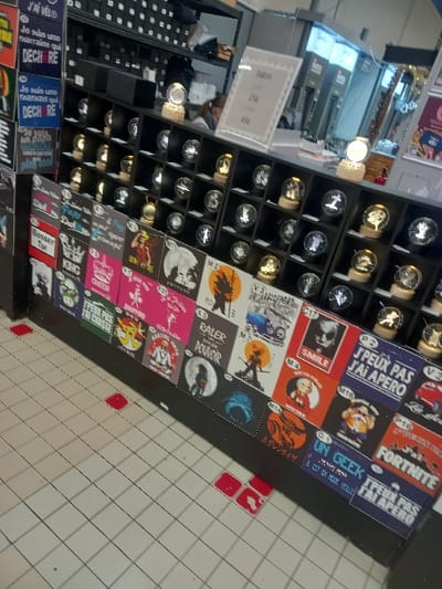 The image displays an indoor retail environment located in Vendin-le-Vieil, France. A prominent black display unit occupies the foreground and midground. Its lower section is covered with a grid of rectangular prints featuring diverse graphic designs and French text, including phrases such as "DECH RE," "Raler Pour Pouvoir," and "FORTNITE." The upper section of this unit showcases multiple rows of small, illuminated crystal globes, each containing a laser-etched 3D design and resting on a light-emitting base, some appearing gold-colored. A white pricing sign, partially visible in the upper right, indicates costs of "25€" and "45€."
In the upper left background, a female individual with dark hair is partially visible behind the display unit, facing away from the camera. Further back on the left, black shelving units are stocked with numerous dark, rectangular boxes. The flooring consists of light-colored square tiles, marked by several scattered small red square adhesive patches. The overall environment is illuminated by artificial lighting, characteristic of a commercial interior. No active interactions are observable in the scene.
