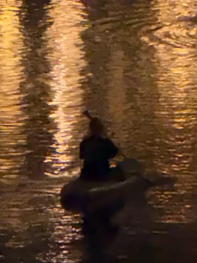A person is in a raft on a body of water, possibly a canal, in Ghent, Belgium. The water reflects streetlights or other artificial light sources, creating a shimmering effect. The person appears to be using a paddle to navigate the raft. The overall scene is dark, suggesting it is nighttime. The raft is a dark color, and the person is silhouetted against the water's reflection. The resolution of the image is low, making it difficult to discern fine details.