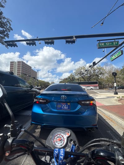 The image depicts a first-person view from a Honda scooter stopped at the intersection of SW 13th Street (also designated Dr. Martin Luther King Jr. Hwy) and SW 8th Street in Gainesville, Florida, United States.

In the immediate foreground, the handlebars of a scooter are visible, along with a circular speedometer displaying an odometer reading of "18554" and a maximum speed of "80" with a redline at "60". A blue "UNIVERSITY OF FLORIDA" lanyard hangs from the ignition keys.

Directly in front, a blue metallic Toyota Camry (license plate "FLORIDA 25DJ" with a University of Florida Gators graphic) is stopped. To the left of the Camry, a dark-colored SUV is also stopped.

Overhead, a traffic light gantry spans the intersection, supporting multiple traffic signals and two green street signs: "SW 13th St Dr. Martin Luther King Jr. Hwy" and "SW 8th". Traffic cameras are mounted on the gantry.

In the background, a large, multi-story brown brick building with numerous windows is visible on the left. To the right, a white passenger bus is seen partially crossing the intersection, with other vehicles beyond it. The weather is clear with a bright blue sky and scattered white cumulus clouds. Large trees, some with bare branches, line the streets. The road features asphalt pavement with white-striped crosswalks and red brick accents. All vehicles appear to be stationary, indicative of being stopped at a traffic light.