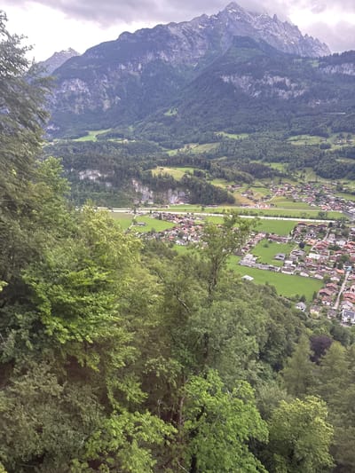 This aerial, medium-distance, eye-level, shot captures a panoramic view of the mountainous landscape and a village nestled in a valley. In the foreground, lush green trees dominate the frame, with their branches extending into the shot. Beyond the trees, a valley unfolds, featuring a small village with red-roofed houses scattered across green fields. A road runs through the valley, connecting the houses. In the background, imposing mountains rise majestically, their peaks shrouded in clouds. The sky is overcast, with dramatic cloud formations adding to the scene's natural beauty. The overall impression is one of a serene and picturesque alpine setting. The location is identified as Hüsen, Switzerland, a charming village renowned for its stunning natural beauty and tranquil atmosphere.
