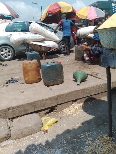 This image depicts a bustling outdoor market scene, characteristic of a street market in a location like Mgbuoshimini, Nigeria. The time of day is bright and sunny, indicated by strong shadows cast on the ground.

In the foreground, the scene unfolds on a concrete pavement and dirt ground. Two large, weathered plastic jerry cans, one orange-brown and the other dark blue-green with a red cap, are prominently placed on the pavement. Beside them lies a green plastic funnel. The ground is littered with scattered light-colored granular material, possibly corn kernels or dried grains, alongside a yellow plastic bag and other debris.

Mid-ground, a man wearing a blue t-shirt and dark shorts is positioned near a silver sedan car, likely a Toyota Yaris. He is attending to a rusty metal wheelbarrow loaded with three large white sacks, presumably containing bulk goods. Partial text "OKE..." is visible on one of the sacks. Behind him and further into the market, several colorful market umbrellas (red, yellow, pink, and white) provide shade for various stalls and vendors. Indistinct figures move amongst these stalls.

To the right, another person, dressed in a patterned blue outfit, is seated on a low stool or container, seemingly engaged in vending or waiting. A large brown barrel or drum and another clear funnel are situated near this person. Another large white sack is also visible in this area.

The scene conveys an atmosphere of active commerce and daily life in an informal market setting, where goods are transported and sold in an open-air environment.
