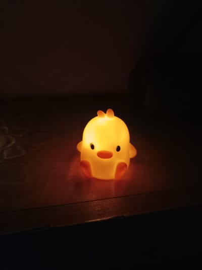 A softly glowing, yellow chick-shaped night light is positioned on a dark wooden surface. The room is dimly lit, casting the rest of the scene in near darkness. There are no people present. The setting's specific location in Cherkovitsa, Bulgaria, is not discernible from the visual information provided.