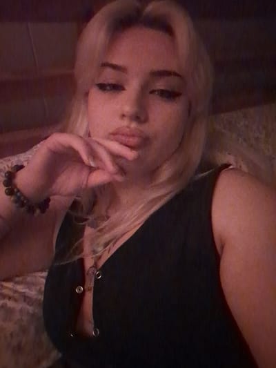 The image displays a close-up portrait of a single individual, a young adult female, positioned in an indoor setting. She has long, blonde hair with bangs, light skin, and visible freckles on her nose and cheeks. Her eyes are accentuated with winged black eyeliner, and her lips are pursed. Her left hand is raised, with fingers resting under her chin. A multi-beaded bracelet, composed of small, dark-colored beads, is worn on her left wrist. She is dressed in a dark-colored, sleeveless top featuring a V-neckline secured with metallic eyelets, some of which are unfastened. An elongated, light-colored pendant on a necklace is visible in her décolletage. The background is dimly lit and indistinct, showing a dark, textured surface behind her left side and a blurred, dark area behind her right side. A light-colored, patterned fabric is visible behind her left shoulder. The lighting is warm and subdued. The precise location within Area D (Elaho/Garibaldi), Canada, is not discernible from the visual information provided in the image.