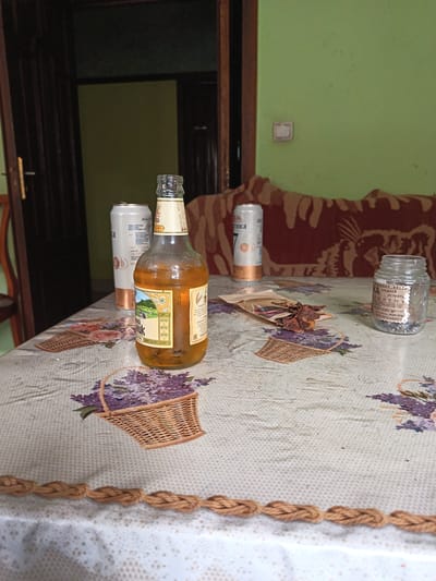 This is a medium-shot, eye-level photograph of a table covered with a patterned tablecloth featuring baskets of lavender. The focus is on the items placed on the table.
To the left, a partially opened doorway reveals a dark interior and a glimpse of a wooden chair. In the background, a light green wall is visible, with a brown and red patterned sofa or chair against it.
On the table, the prominent object is a glass bottle of amber-colored liquid, likely beer or a similar beverage, with a label that includes imagery of a landscape. Next to it, two white aluminum cans are positioned, one with Cyrillic text. In front of the cans, a small, open brown paper packet contains pieces of dried, dark-colored jerky or meat. To the right of the jerky is a small, clear glass jar with a white label that also has Cyrillic text. The label on the jar appears to indicate a date of "25.09.2024".
The lighting suggests an indoor setting, possibly during the day, with ambient light illuminating the scene. The overall impression is that of a casual setting, perhaps a home, with refreshments and snacks laid out. The visible text includes brand names and descriptions, predominantly in Russian, such as "ЭКСПОРТНО", "СТАРОГКОЙ", and "СРЕДНЕЙ ОСТРОТЬ".