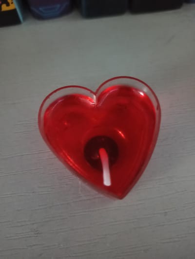 An overhead, close-up shot shows a red, heart-shaped candle with a white wick inside a clear, heart-shaped casing. The candle is placed on a light gray, textured surface. The background is out of focus and shows dark objects. The scene is located in Promyshlennaya, Russia.