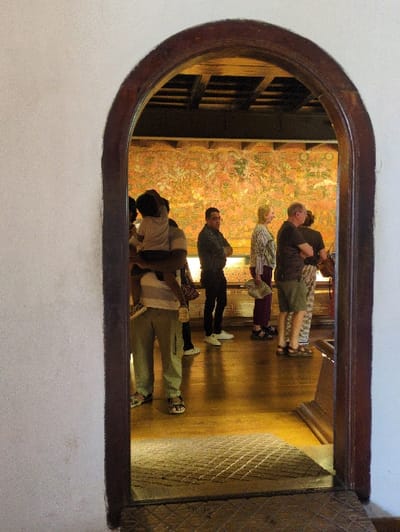 The image displays an interior view framed by a dark brown arched wooden doorway, likely situated within the Mattancherry Palace in Fort Kochi, Kochi, India. The foreground shows a light-colored, textured wall. Beyond the archway, an interior room features a reflective, polished wooden floor and a dark ceiling with exposed wooden beams. The far wall is dominated by a large, intricately detailed mural, exhibiting a palette of warm orange, red, and yellow with green and white accents, consistent with traditional Kerala mural art depicting figures and scenes.
Approximately six individuals are present in the room, all oriented towards and observing the mural. On the left, partially within the doorway's frame, a person wearing light trousers and a striped top carries a small child on their shoulders; the child faces the mural, reaching upwards with one arm. To the right, a man in a dark jacket stands with arms crossed. Further right, a woman with light hair, wearing a patterned top, holds a folded document. Beside her, a man in a dark t-shirt and light shorts stands. Additional figures are partially visible further right. The room is illuminated, with the mural appearing distinctly bright.