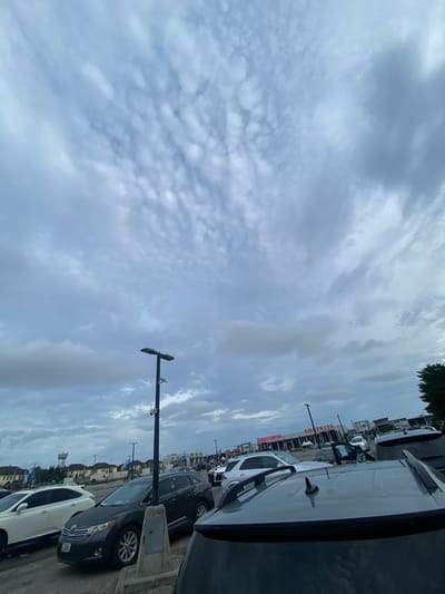 The image shows a parking lot with cars parked along the edge, adjacent to a Shoprite supermarket, potentially in Sangotedo, Nigeria. Overcast skies dominate the view. Streetlights are visible alongside parked cars.