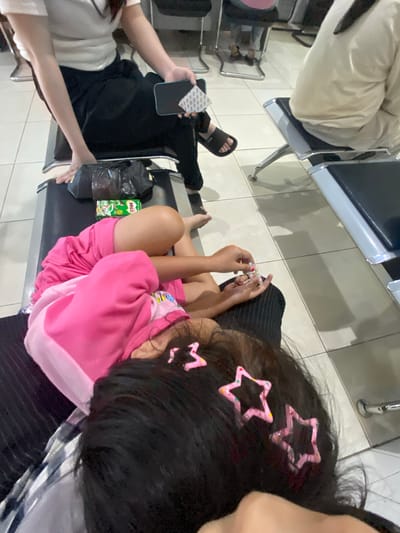 In Ta', Indonesia, at a waiting area, a young girl in a pink sweatshirt lies down on a bench, her head resting on someone's lap. She is adorned with star-shaped hair clips. Her nails appear to be painted with multiple colors. Next to her, a green Milo box and a black plastic bag are placed on the bench. Another person, dressed in black pants and a white t-shirt, sits beside her, holding a light blue smartphone with a sticker attached. They are wearing black slide sandals. The floor is tiled.