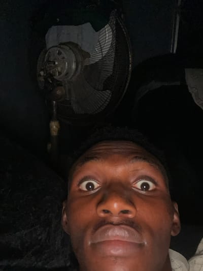 A close-up, waist-up selfie is taken by a young Black male who is lying down and looking up at the camera. The man is in a dark room, likely his bedroom, with a standing fan visible behind him. He is wearing a white shirt with a textured pattern around the neckline. His eyes are wide open, and his mouth is slightly parted. The lighting suggests it may be nighttime or a dimly lit room. The image was taken in Akowonjo, Nigeria.