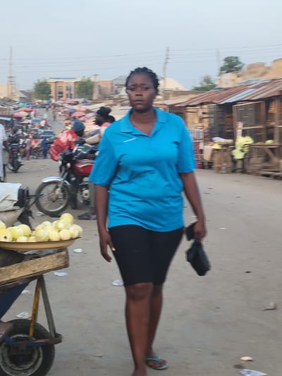 An outdoor, daylight shot depicts a woman in Kwamba, Nigeria, standing near her merchandise on a busy street. The woman, wearing a blue shirt and black skirt, stands next to a handcart filled with yellow fruits, possibly mangoes or pears, ready for sale. Behind her, a red motorcycle is parked amidst the pedestrian traffic. The street scene is filled with people and various stalls, indicating a bustling marketplace. In the background, buildings and power lines are visible under a hazy sky.