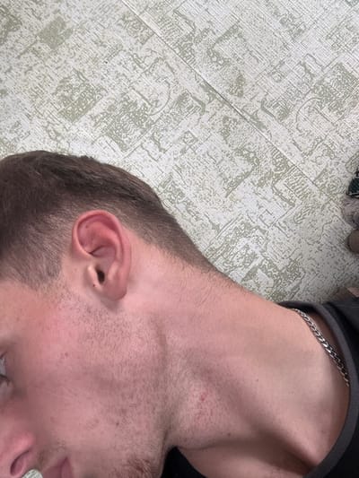 A close-up, angled photograph taken in the city of Durgena, Georgia, depicts the left side of a male individual's head, neck, and upper chest. The individual has short, dark brown hair with a faded cut above the ear. The left ear is fully visible, exhibiting no apparent anomalies. Light stubble is present on the cheek, jawline, and chin. The skin is fair, with minor redness on the lower neck and cheek, and a small red blemish is visible on the left side of the neck/upper chest. A silver-colored metal chain necklace is worn around the neck, resting over a dark-colored garment with a gray trim. The background consists of a light-colored surface with a repeating, pale green or light grey abstract geometric pattern, consistent with wallpaper or a decorative wall panel. A diagonal seam in this surface is visible in the upper half of the image.