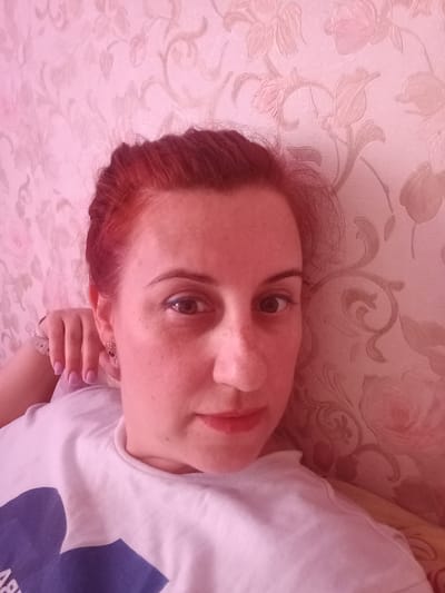 A selfie shows a fair-skinned woman with red hair and freckles against a patterned pink wallpaper with floral and scroll designs. The woman is wearing a white t-shirt with a dark blue graphic on the front. Her left arm is raised, with her hand near her left ear. The image was taken in Serpukhov, Russia.