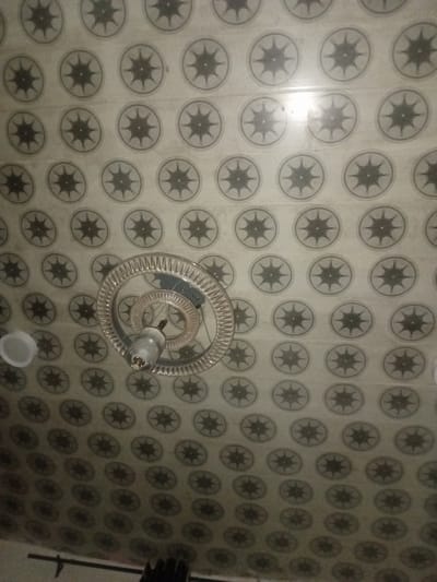 This image captures the interior ceiling of a room in Ogbogbo, Nigeria, viewed from a low-angle perspective looking upwards. The ceiling is uniformly covered with light-toned rectangular panels, likely PVC or similar material, arranged in a grid. Each panel features a dark, repeating circular motif containing a stylized eight-pointed star or sunburst design, set against an off-white or light grey background.
A modern chandelier is suspended centrally from the ceiling. It is constructed from multiple concentric, clear, possibly acrylic or glass, rings, with the outermost ring displaying a ribbed or textured edge. A single, elongated light fixture is mounted at the core of these rings, enclosed in a clear or frosted shade, providing artificial illumination. Reflections from this light source are visible on the surrounding ceiling panels and the polished surfaces of the chandelier. To the left of the chandelier, a partial white, inverted dome-shaped object, possibly a light cover or vent, is visible. The very bottom edge of the image shows indistinct dark architectural elements, likely part of a wall or another fixture below the ceiling line. No individuals are present in the scene.