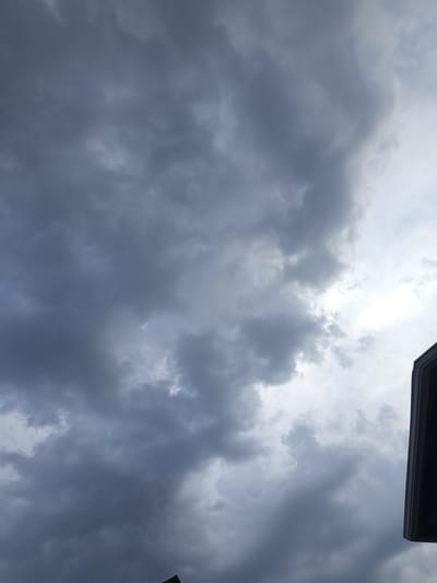 The image captures a dramatic sky filled with dark, heavy storm clouds. The clouds are dense and varied in texture, with some areas appearing lighter and more diffused, suggesting the presence of sunlight trying to break through. The overall tone is somber and foreboding, indicative of impending or ongoing bad weather. In the lower right corner, the edge of a dark, angular object, possibly a building or a sign, is visible, providing a slight sense of grounding to the vast expanse of the sky. The scene appears to be an outdoor setting, likely during the daytime, given the brightness of the sky. The mood evoked is one of an overcast, possibly rainy or stormy day. No people or discernible text are present in the image. The location context of Жаны-Кыштак, Kyrgyzstan, places this potentially turbulent weather event in a specific geographical region, though there are no direct visual cues within the image to confirm this location.