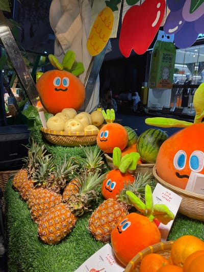 The image is of a fruit stand in Hangzhou, China (30.263, 120.156). The stand is decorated with cartoon orange characters, with the main character being a smiling orange with large blue eyes and a small mouth. The orange character is surrounded by pineapples and watermelons. The fruit stand is situated on a green artificial grass base. In the background, there is a person in a black shirt standing behind a table. The scene takes place in a well-lit, brightly colored environment. The fruit stand is located in a retail setting, possibly within a larger market or shopping mall.