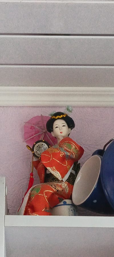 A detailed, forensic, matter-of-fact description of the scene is as follows:
The image depicts a decorative doll, likely a Kokeshi or a similar traditional Japanese doll, positioned on a shelf. The doll is adorned in an elaborate, brightly colored kimono, predominantly red with gold and green patterns. It is holding a small pink umbrella and a drum-like object with a gold handle. The doll has black hair styled with a gold band and small green decorative elements.
The doll is situated against a textured pink background. To the right of the doll, on the same shelf, are two large blue cups, one of which is tilted, and a smaller blue and white patterned ceramic pot or jar. The shelf itself is white and appears to be part of a larger shelving unit, indicated by a white vertical edge on the left side of the frame. Above the doll and shelf, a white decorative molding is visible, against a background of horizontally lined gray material.
There are no people present in the image, nor any indications of specific activities or interactions. The environment appears to be indoors, likely a display shelf in a home or a shop.
Regarding the location in Tikhvin, Russia, there is absolutely no visual information within the image that could suggest or confirm this location. The subject matter, a traditional Japanese doll, is culturally specific to Japan and does not inherently connect to Tikhvin or any other location in Russia without further context.
