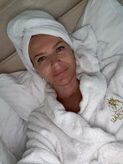A woman with a white towel wrapped around her head and a white terrycloth robe is lying in bed. She is looking at the camera and smiling. The robe has a logo on the left side. The background is a white pillow. The image does not provide any information about the location in the city of Barangol, Russia.