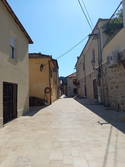 The image depicts a narrow, sun-drenched street in Trebinje, Bosnia and Herzegovina. The street is paved with light-colored stone tiles. On the left side of the street, a light yellow building with a window covered by a roller shutter and a barred entrance dominates the foreground. Its shadow stretches across the pavement. Further down the street, more buildings are visible, including a yellow structure with a red-tiled roof and various coiled cables attached to its exterior. The right side of the street features a building with a stone facade, an air conditioning unit, and an outdoor light fixture. Overhead, power lines crisscross the clear blue sky. The scene conveys a sense of quiet, Mediterranean ambiance.