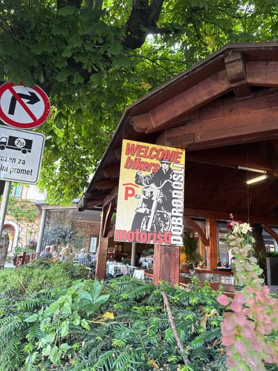 The image shows a sign reading "WELCOME bikers motoristi DOBRODOSLI" mounted on a wooden structure, possibly a covered patio, in Bled, Slovenia, located at the coordinates 46.368 latitude and 14.113 longitude. The sign features a man riding a motorcycle. The sign is partially obscured by green foliage, and is located on a street corner where other road signs are visible. Several people are sitting at tables in the background. The scene is a warm and inviting environment, with the green foliage suggesting a setting in nature.