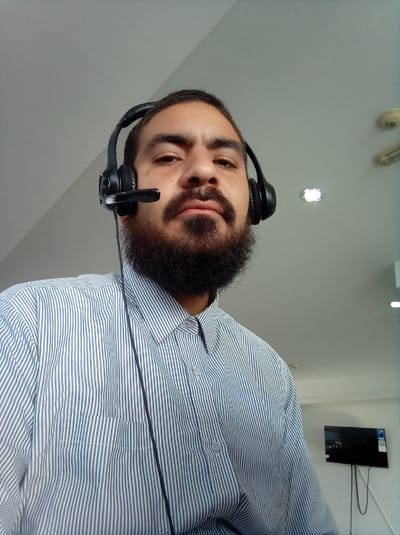 The image shows a man from the chest up. He has a dark beard and mustache. He is wearing a blue and white striped button-down shirt and a black headset with a microphone. The background is a white ceiling with recessed lighting and a TV mounted on a white wall. The location is presumably in an office or similar setting in Parroquia Guatire, Venezuela.