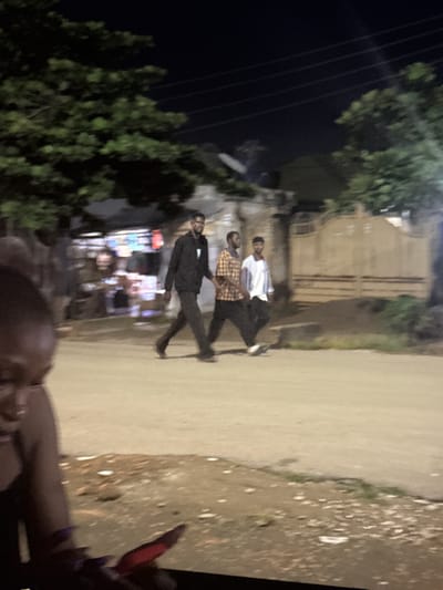 The image depicts a nighttime street scene in Abuja, Nigeria. Three men are walking on a paved road. The man on the left wears a black hooded jacket and dark pants. The middle man is wearing a plaid shirt and dark pants. The man on the right is wearing a light-colored long-sleeved shirt and dark pants. In the background, there are trees, buildings, and a fence. The road appears to be dusty and uneven, with some debris scattered along the edges. The lighting suggests artificial illumination from streetlights or nearby establishments. A portion of what appears to be a person's head and shoulder, possibly a woman, is visible in the foreground on the left side of the image, out of focus.