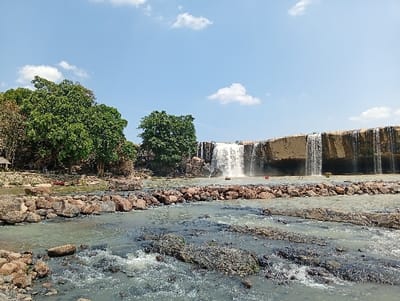 The image presents a wide view of a waterfall and river landscape situated in Đức Lập, Vietnam, under a clear blue sky adorned with scattered white cumulus clouds. A broad waterfall dominates the right half of the midground, cascading in multiple distinct streams from a wide, stratified, light-brown rock cliff into the river below. The river in the foreground and midground features greenish-brown water, with numerous dark, irregular rocks and small rapids visible. A deliberate formation of large, reddish-brown rocks creates a linear barrier or pathway extending across the river in the midground. The left bank is densely covered with vibrant green trees and foliage, partially obscuring small, light-colored structures, possibly gazebos or shelters. A red object, appearing to be a small boat or platform, is partially visible near the water's edge on the left bank. In the water directly below the main waterfall, a small, dark object and a red sign with illegible text are present. No individuals are explicitly visible. The scene is brightly illuminated by daylight.