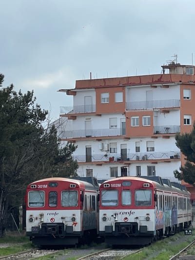 The image displays two stationary Renfe commuter trains on parallel railway tracks in Buñol, Spain, positioned in front of a multi-story residential building under an overcast sky. The foreground features two red and white train cars. The train on the left is numbered "301M" on its destination board, while the train on the right is numbered "502M." Both units exhibit the "renfe" logo on their lower front sections. The side of the 502M train is adorned with multicolored graffiti, and the side of the 301M appears weathered with minor graffiti. Green vegetation, including grass and weeds, grows between the railway sleepers and tracks.
Behind the trains and partially obscured by sparse trees on the left, a six-story residential building is prominent. The building features predominantly white facades with horizontal bands of terracotta-orange trim, particularly on the upper levels and around balconies. Multiple balconies with white metal railings extend from each floor. Numerous rectangular windows, some with closed shutters, are visible across the building's face. Exterior air conditioning units are mounted below some windows. The flat roof of the building hosts an antenna array and a small, elevated utility structure. The sky above is uniformly grey and overcast. Several small, pink, irregularly shaped objects are visible in the upper central portion of the sky.
