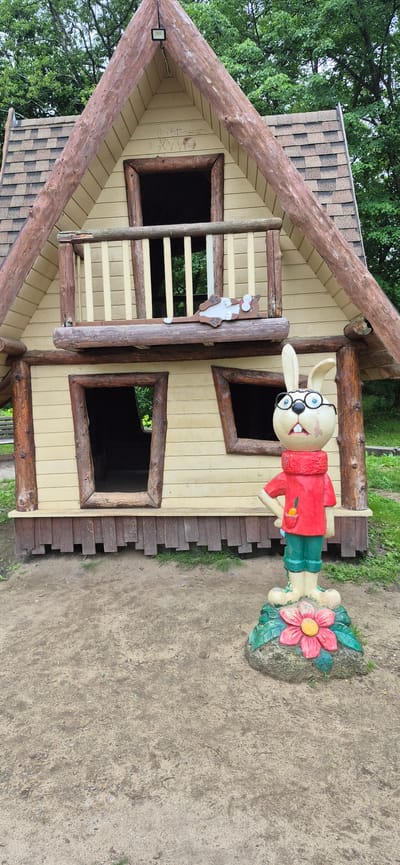 The image shows a whimsical wooden playhouse designed to resemble a small cabin. The playhouse has a steeply pitched roof covered with shingles and is constructed with light-colored siding and natural wood accents, including log beams and window frames. There are several openings that serve as windows and a door, some with wooden shutters. A small balcony is visible on the upper level, with a ladder leading up to it. A decorative cutout of a brown and white cat hangs from a wooden post next to the ladder.
To the right of the playhouse, a brightly colored, anthropomorphic statue of a rabbit stands. The rabbit is wearing glasses, a red sweater, and green pants. It is holding a small orange carrot in its hand and stands beside a sculpted pink flower with a yellow center, which is attached to a rock. The ground around the playhouse and statue is covered in dirt and scattered with small stones and patches of grass. Lush green trees and foliage surround the play area. The lighting suggests it is daytime, with diffused light indicating potentially overcast skies or heavy tree cover. The scene appears to be an outdoor playground or park, possibly in a rural or semi-rural setting. There are no visible text elements in the image.